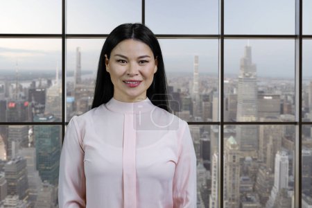 Foto de Pretty young asian business woman standing in office. Smiling korean brunette. Windows with cityscape background. - Imagen libre de derechos