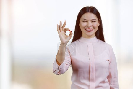 Foto de Portrait of happy asian woman showing okay gesture. Blurred office windows background. - Imagen libre de derechos