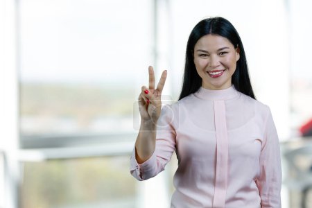 Téléchargez les photos : Portrait of happy asian woman showing victory sign. Blurred office windows background. - en image libre de droit