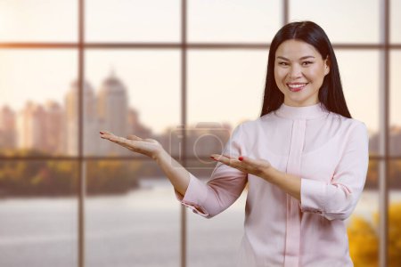 Foto de Young smiling asian woman holding copy space on her palms. Checkered windows background. - Imagen libre de derechos