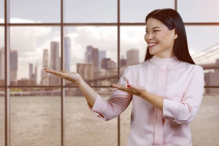 Téléchargez les photos : Portrait of young asian woman showing copy space with both hands. Windows with cityscape on the background. - en image libre de droit