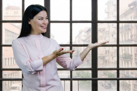 Téléchargez les photos : Portrait of young asian woman advertising copy space with both hands. Checkered windows with cityscape on the background. - en image libre de droit