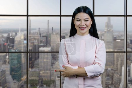 Téléchargez les photos : Portrait of young smiling asian woman pointing at copy space. Checkered windows with cityscape on the background. - en image libre de droit