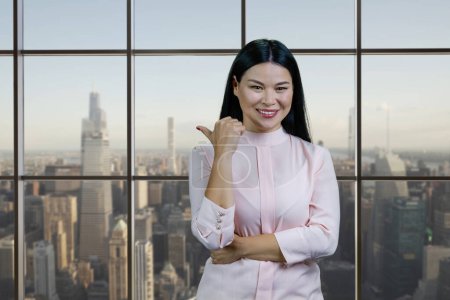 Téléchargez les photos : Portrait of young smiling asian woman pointing back. Checkered windows with cityscape on the background. - en image libre de droit