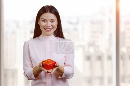 Téléchargez les photos : Smiling asian woman giving red gift box. Blurred windows background. - en image libre de droit