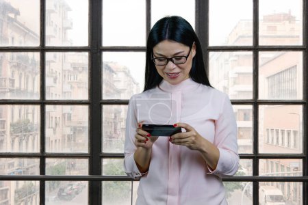 Foto de Young asian businesswoman watching a video on her smartphone device. Checkered windows background. - Imagen libre de derechos