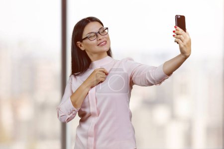 Photo for Young korean businesswoman in glasses is taking a selfie. Blurred windows background. - Royalty Free Image