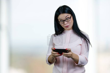 Foto de Asian businesswoman is watching something on her smartphone with eyes wide open. Abstract blurred background. - Imagen libre de derechos