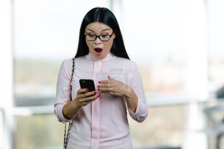 Téléchargez les photos : Portrait of extremely surprised asian woman looking at her smartphone. Mouth wide open. Blurred abstract indoor background. - en image libre de droit