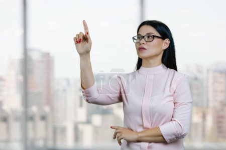 Photo for Young asian business woman is touching invisible virtual screen by her finger. Blurred cityscape background. - Royalty Free Image