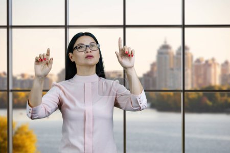 Photo for Young asian woman is interacting with invisible virtual screen by both hands. Checkered windows with cityscape background. - Royalty Free Image