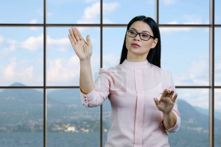 Photo for Young asian businesswoman is swiping something on imaginary virtual screen by her hand. Checkered windows with mountainscape background. - Royalty Free Image