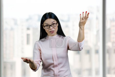 Foto de Portrait of expressive asian business woman is gesturing actively. Blurred windows background. - Imagen libre de derechos