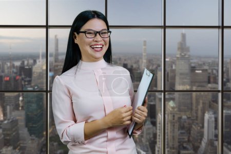 Téléchargez les photos : Smart young asian businesswoman with clipboard is laughing. Checkered windows background with cityscape view. - en image libre de droit