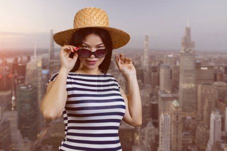Téléchargez les photos : Portrait of attractive young asian woman in sunglasses and straw hat. Big cityscape background. - en image libre de droit
