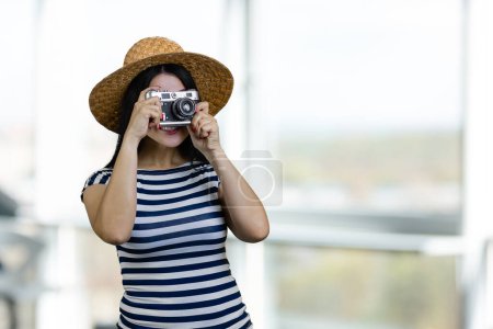 Photo for Young tourist woman in straw hat is taking a photo on vintage photo camera. Indoor windows background. - Royalty Free Image