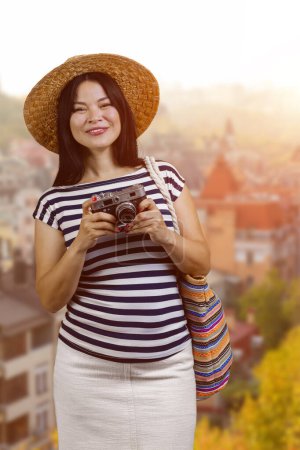 Foto de Portrait of asian female tourist in straw hat is holding a vintage photo camera. Vertical shot blurred townscape background. - Imagen libre de derechos