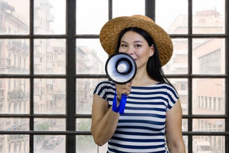 Téléchargez les photos : Young korean woman in straw hat is giving an announcement in megaphone. Checkered indoor windows background. - en image libre de droit