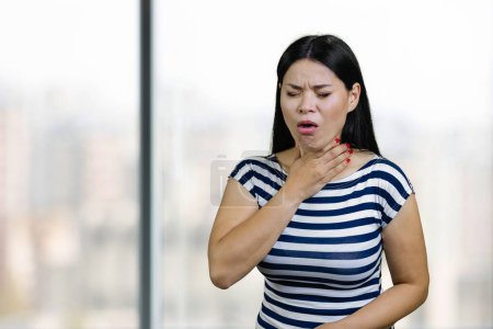 Téléchargez les photos : Young asian woman is coughing and touching her throat. Office windows background. - en image libre de droit