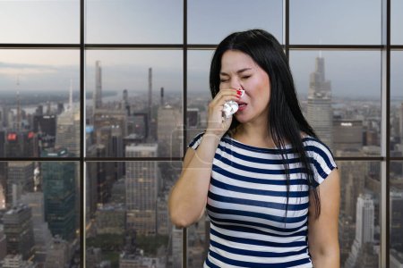 Téléchargez les photos : Portrait of a young asian woman is sneezing in handkerchief. Checkered windows background with cityscape view. - en image libre de droit