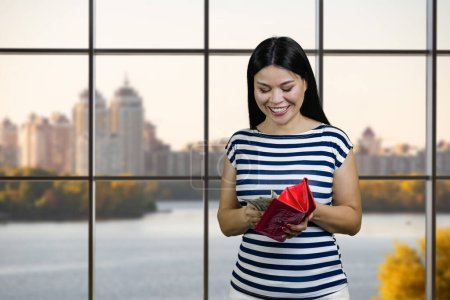 Foto de Happy asian woman is taking money from the red wallet. Checkered windows with cityscape view on background. - Imagen libre de derechos