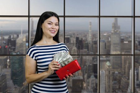 Foto de Young asian woman is taking money from the red wallet. Checkered windows with cityscape on background. - Imagen libre de derechos