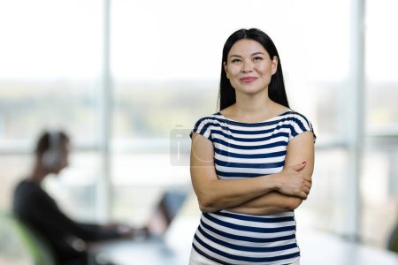 Téléchargez les photos : Portrait of young asian casual-dressed woman with folded arms is looking up. Blurred office enviroment background. - en image libre de droit