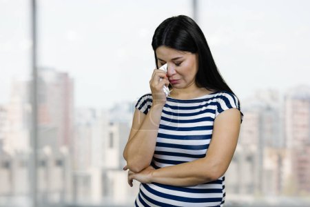 Photo for Young asian woman is crying and wiping her tears. Sad upset korean female standing indoors. Windows with cityscape on background. - Royalty Free Image