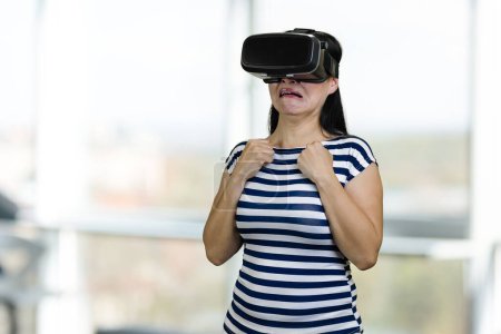 Foto de Young woman is afraid of something in virtual reality glasses. Blurred windows background. - Imagen libre de derechos
