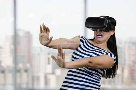 Foto de Scared woman in vr headset stretching her arms saying stay away from me. Blurred windows background. - Imagen libre de derechos