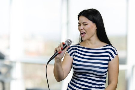 Téléchargez les photos : Young brunette asian woman is singing a song with microphone. Blurred bright background. - en image libre de droit