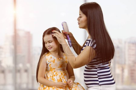 Foto de Young brunette mother is combing hair of her little daughter. Blurred window indoor background. - Imagen libre de derechos