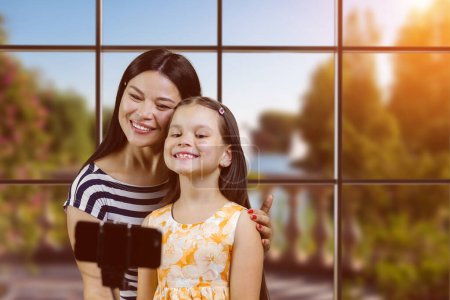 Téléchargez les photos : Mother and daughter are making a selfie on smartphone using the stick. Checkered windows with city park view. - en image libre de droit