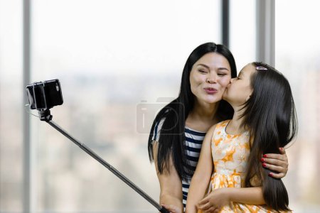 Téléchargez les photos : Mother and her daughter are making a selfie on smartphone using the stick. Little girl kissing her mom for photo. Blurred windows background. - en image libre de droit