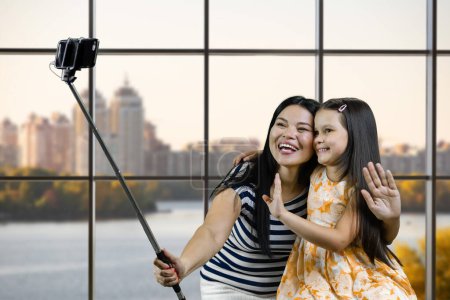 Téléchargez les photos : Mom and her daughter are making a common photo on smartphone using selfie stick. Blurred cityscape background. - en image libre de droit