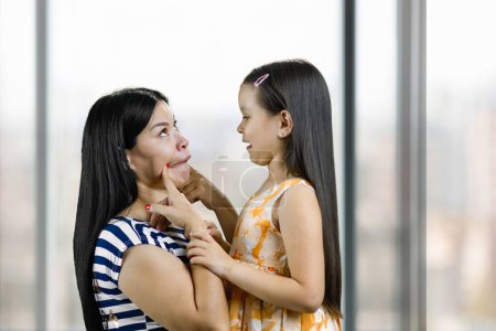 Photo for Mother making funny face to her little daughter. Woman pulling her cheeks. Blurred indoor windows background. - Royalty Free Image