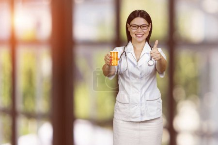 Foto de Young female doctor is showing medicine can and giving thumb up. Advertise and promotion. Blurred checkered background. - Imagen libre de derechos