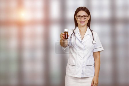 Foto de Happy young female asian doctor is advertising medicine bottle. Blurred checkered windows background. - Imagen libre de derechos
