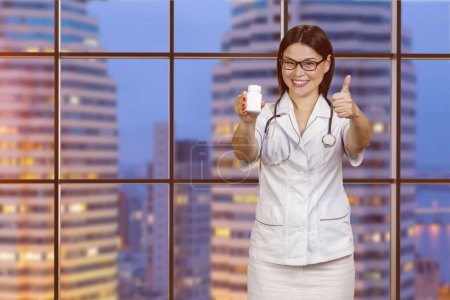 Foto de Young female doctor in glasses is showing medicine bottle and giving thumb up. Checkered windows with night cityscape view. - Imagen libre de derechos