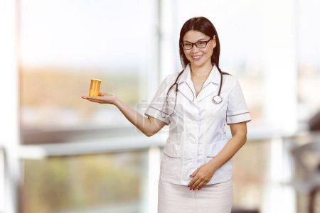 Foto de Young female doctor in glasses is showing orange medicine bottle on her palm. Blurred window background. - Imagen libre de derechos