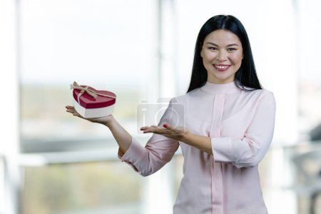 Foto de Cheerful asian woman showing heart-shaped gift box. Standing indoors in office against windows. - Imagen libre de derechos