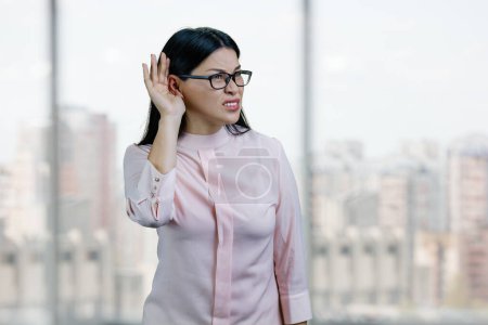 Foto de Young asian business woman try to hear you out.Overhear listening sound to gossip. Checkered windows and cityscape background. - Imagen libre de derechos
