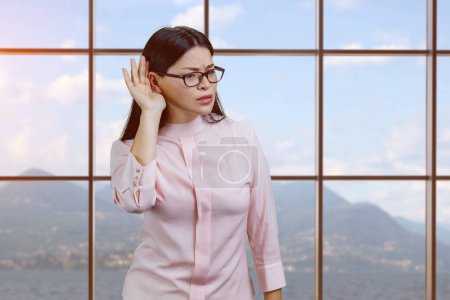 Foto de Young asian businesswoman struggles hard to hear you. Checkered window and landscape background. - Imagen libre de derechos
