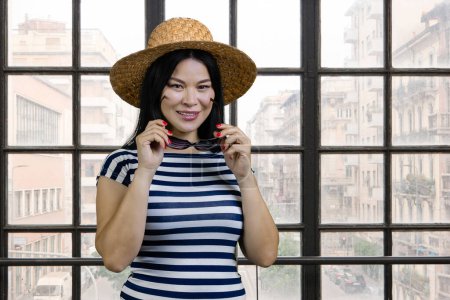 Téléchargez les photos : Portrait of a young asian woman wearing straw hat and sunglasses. Checkered windows background with cityscape view. - en image libre de droit