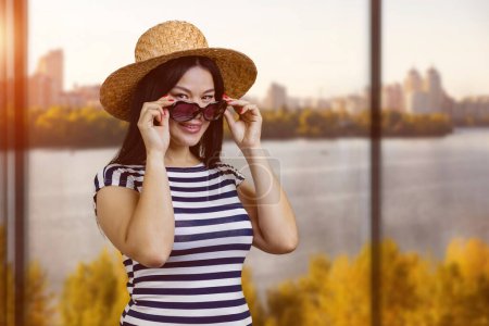 Téléchargez les photos : Portrait of a young asian woman in straw hat wearing sunglasses. Window with autumn landscape background. - en image libre de droit