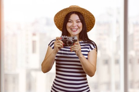 Foto de Portrait of a young smiling tourist asian woman with vintage photo camera. Blurred windows background. - Imagen libre de derechos