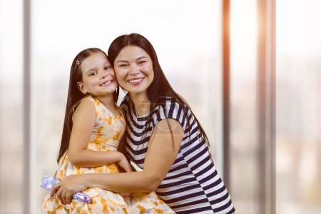 Téléchargez les photos : Portrait of a happy asian woman with her adopted caucasian little daughter. Cute little girl with her mother indoors. Blurred windows background. - en image libre de droit