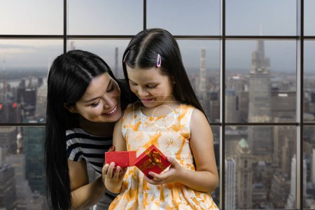 Foto de Happy little girl is opening her birthday gift box from her loving mother. Checkered windows background with cityscape view. - Imagen libre de derechos