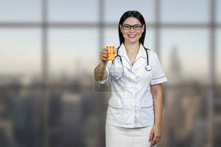 Téléchargez les photos : Portrait of a young female asian doctor is showing medicine can. Smiling cheerful brunette physician with glasses and stethoscope. - en image libre de droit