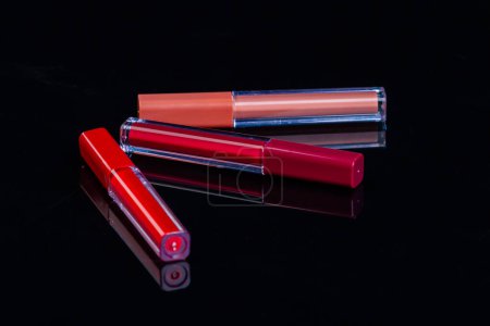 Téléchargez les photos : Three lip gloss sticks on reflective surface. Cosmetic makeup tools on black background. - en image libre de droit
